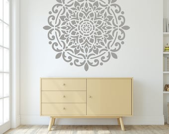 May include: A gray mandala wall stencil with a decorative floral design. The stencil is on a white wall above a light beige dresser with three drawers.