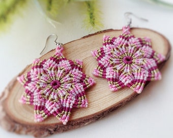 May include: A pair of pink and gold star-shaped earrings with a mandala design. The earrings are made using a macrame technique and are hanging from silver hooks.