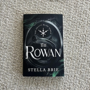 May include: A black and green book cover with the title "The Rowan" and the author "Stella Brie". The cover features a circular design with the text "The Killian Blade Series" and a stylized sword.