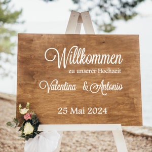 May include: A wooden sign with white text on a white easel. The sign says "Willkommen zu unserer Hochzeit Valentina & Antonio 25 Mai 2024".