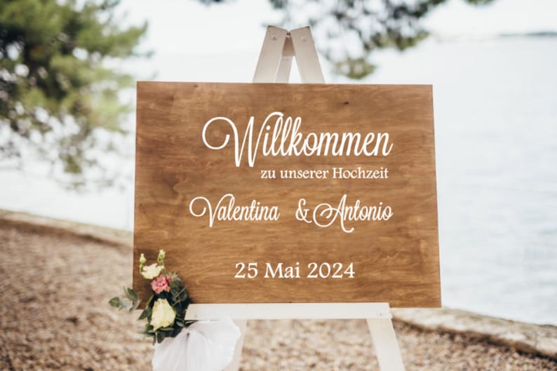 May include: A wooden sign with white text on a white easel. The sign says "Willkommen zu unserer Hochzeit Valentina & Antonio 25 Mai 2024".