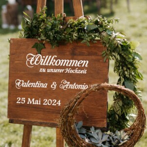 May include: A wooden sign with the text "Willkommen zu unserer Hochzeit" (Welcome to our wedding) and the names "Valentina & Antonio" and the date "25 Mai 2024". The sign is decorated with greenery and is propped up on an easel.