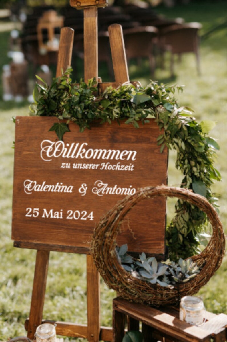 May include: A wooden sign with the text "Willkommen zu unserer Hochzeit" (Welcome to our wedding) and the names "Valentina & Antonio" and the date "25 Mai 2024". The sign is decorated with greenery and is propped up on an easel.