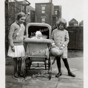 Puede incluir: Una fotografía en blanco y negro de tres niños. Dos niñas están de pie en una acera, una sosteniendo un cochecito de mimbre con un bebé dentro. La otra niña sostiene un sombrero.
