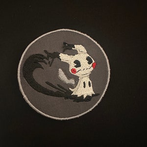 May include: A gray embroidered patch with a white ghost-like creature with red cheeks and black eyes. The creature is surrounded by a black shadow.