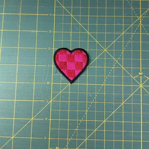 May include: A heart-shaped patch with a black border. The heart is filled with a checkered pattern of pink and red squares. The patch is on a green cutting mat.