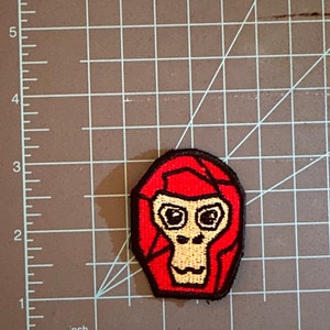 Könnte beinhalten: Roter gestickter Aufnäher mit einem Affengesicht, schwarzen Umrissen und gelben Augen.