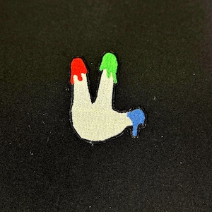 May include: A white embroidered patch shaped like a hand with three fingers. The fingers are tipped with red, green, and blue paint.