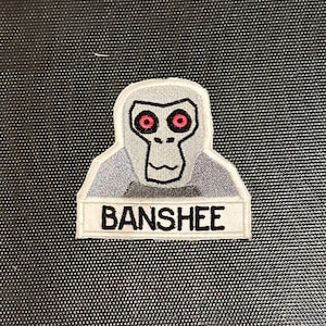 May include: A white embroidered patch with a cartoon gray creature with red eyes and the word "BANSHEE" in black text.