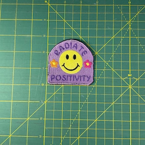 Puede incluir: Un parche morado con una carita sonriente amarilla y las palabras "RADIATE POSITIVITY". El parche tiene una parte superior curva y está decorado con pequeños diseños florales a ambos lados de la carita sonriente.