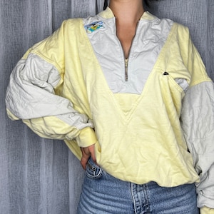Könnte beinhalten: Ein Vintage-Pullover-Sweatshirt in Gelb und Grau mit einem Viertel-Reißverschluss am Halsausschnitt. Das Sweatshirt hat gesteppte graue Ärmel und ein graues Panel auf der Brust. Ein kleiner Aufnäher ist auf der oberen Brust sichtbar. Das Kleidungsstück wird mit Jeans getragen.