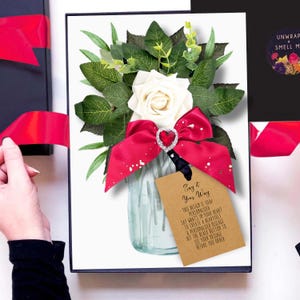 May include: A gift box with a floral arrangement featuring a white rose, green leaves, and a red bow with a heart-shaped embellishment. A tag reads "Say it Your Way". The box is black with a red ribbon.