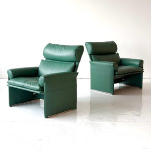 May include: A pair of emerald green leather armchairs featuring high backrests, integrated headrests, and solid side panels. Each chair has a cushioned seat and back, supported by a low-profile base with visible metallic accents at the bottom edges.