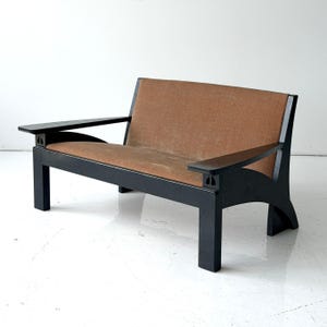 May include: A two-seater sofa featuring a black wooden frame with curved armrests and a solid back support. The seat and backrest are upholstered in a textured, light brown fabric. The structure includes four sturdy legs and open side panels.