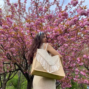 May include: A woman in a white dress is standing in front of a cherry blossom tree. She is carrying a large, beige tote bag with white fringe.