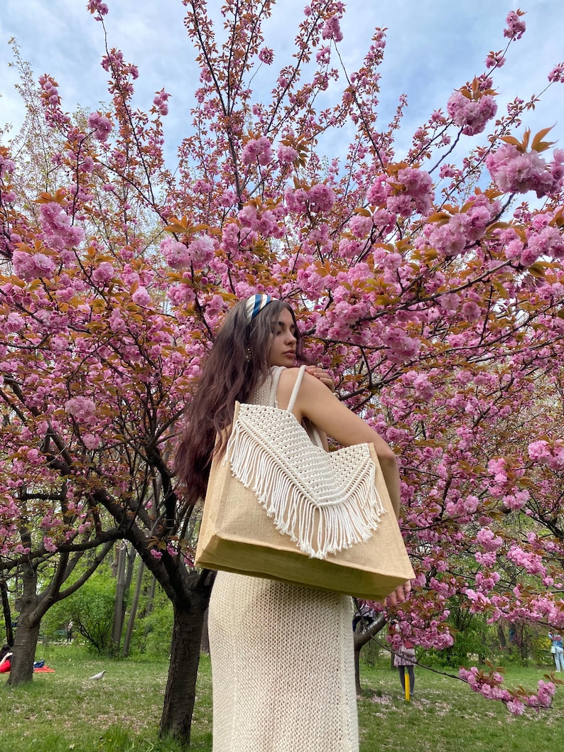 May include: A woman in a white dress is standing in front of a cherry blossom tree. She is carrying a large, beige tote bag with white fringe.