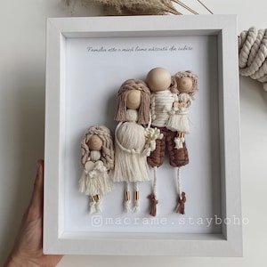May include: A white framed picture with a quote in Romanian that translates to "Family is a small world born of love." The picture features a family of three made of yarn and wood. The family is made of a mother, father, and child. The mother and child are wearing white yarn dresses, and the father is wearing brown yarn trousers. The family is holding hands and smiling.