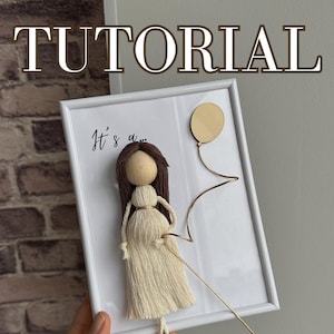 Op de afbeelding: Een wit frame met een witte achtergrond en een bruine garenpop met bruin haar. De pop houdt een gouden touwtje vast dat aan een houten cirkel is bevestigd. De tekst "It's a..." staat op het frame.