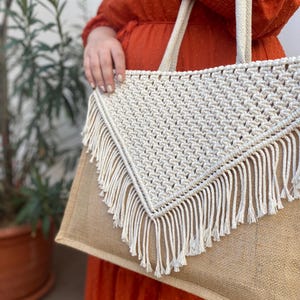 May include: A large, beige tote bag with a white crocheted triangle flap and fringe detail. The bag has a brown jute base and is made of natural materials.