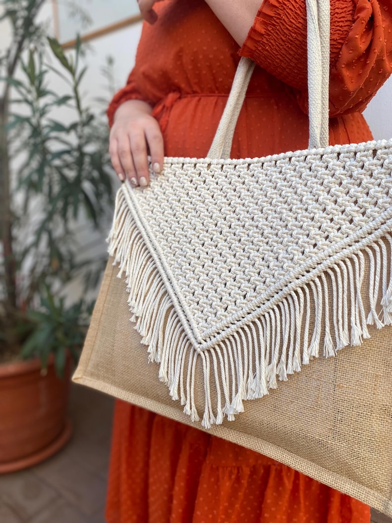 May include: A large, beige tote bag with a white crocheted triangle flap and fringe detail. The bag has a brown jute base and is made of natural materials.