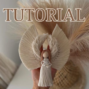 Peut inclure: Un ange en macramé blanc avec une tête en fil rose et un pompon blanc. L'ange est tenu dans une main sur un fond d'herbe de pampa séchée. Le texte "TUTORIAL" est affiché en haut de l'image.