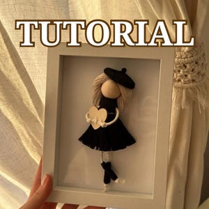 Puede incluir: Una muñeca hecha a mano enmarcada, con un vestido negro, boina y un corazón de madera. La palabra "TUTORIAL" se muestra en marrón en la parte superior del marco. La muñeca está hecha de hilo y otros materiales.
