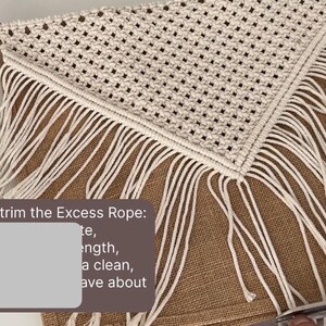 May include: Step number 8, trim the Excess Rope: te, length, a clean, ave about. A close-up of a person trimming the excess rope from a white macrame wall hanging with a fringe. The macrame is hanging on a brown burlap fabric.