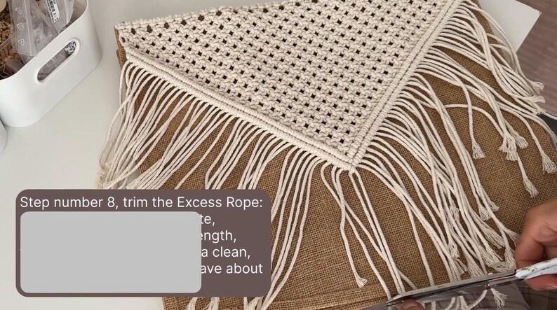 May include: Step number 8, trim the Excess Rope: te, length, a clean, ave about. A close-up of a person trimming the excess rope from a white macrame wall hanging with a fringe. The macrame is hanging on a brown burlap fabric.