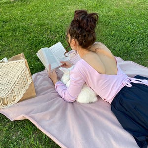 May include: A woman wearing a pink long-sleeved shirt with a low back and a black skirt is sitting on a pink blanket in a grassy area. She is reading a book and holding a white rabbit.