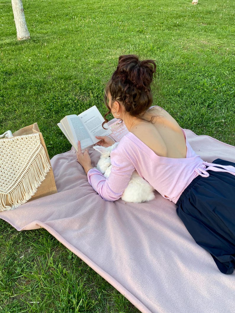 May include: A woman wearing a pink long-sleeved shirt with a low back and a black skirt is sitting on a pink blanket in a grassy area. She is reading a book and holding a white rabbit.