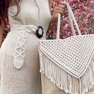 May include: A white crocheted tote bag with a fringe trim and a brown jute base. The bag has a triangular shape with a macrame design.