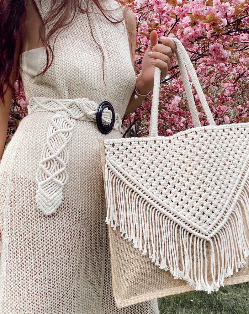 May include: A white crocheted tote bag with a fringe trim and a brown jute base. The bag has a triangular shape with a macrame design.