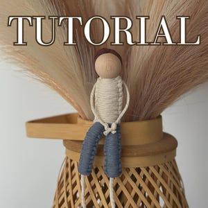 May include: A handmade doll with a wooden head and a brown hat, sitting on a woven basket. The doll is made of white string and has blue trousers. The doll is sitting on a woven basket with a brown rim.
