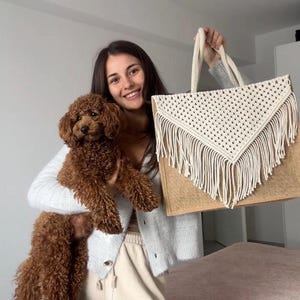 May include: A woman holding a brown and white crocheted tote bag with fringe. She is also holding a brown poodle.