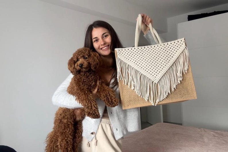 May include: A woman holding a brown and white crocheted tote bag with fringe. She is also holding a brown poodle.