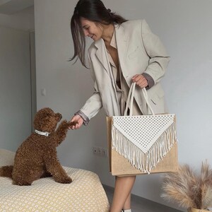May include: A woman in a beige blazer holds a large white crocheted tote bag with fringe. The bag is made of natural jute fabric. A brown dog sits on a yellow and white patterned bedspread.