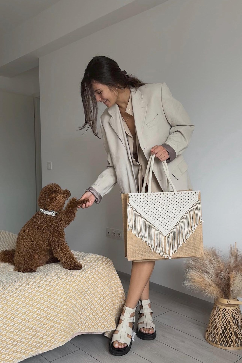 May include: A woman in a beige blazer holds a large white crocheted tote bag with fringe. The bag is made of natural jute fabric. A brown dog sits on a yellow and white patterned bedspread.