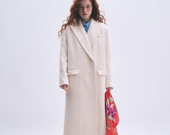Pode incluir: Uma mulher vestindo um casaco branco comprido com uma echarpe floral vermelha. Ela está em pé em um fundo branco.
