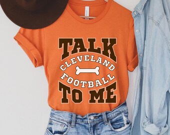 Puede incluir: Camiseta naranja con texto marrón que dice "Talk Cleveland Football To Me" con un gráfico de hueso blanco.