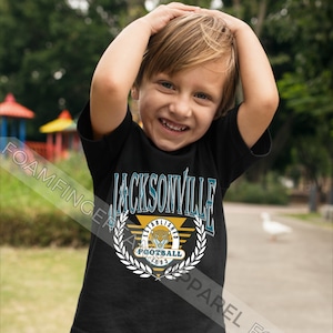 May include: A young child wearing a black t-shirt with a teal and gold Jacksonville Jaguars logo. The logo features a jaguar head, the words "Established 1993", and "Football".