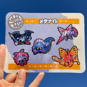 May include: A sheet of stickers featuring various characters from the video game series Kirby. The stickers are on a white background with a glittery texture. The text "STAR DROPS" and "メタナイト" are printed on the sheet.
