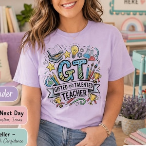 May include: A lavender short-sleeve t-shirt featuring a colorful graphic print with the text GT GIFTED AND TALENTED TEACHER, surrounded by illustrations of books, a lightbulb, beakers, pencils, paintbrushes, and gold trophies.