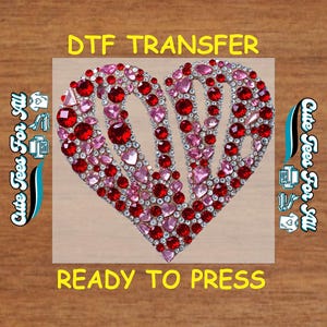 May include: DTF transfer featuring a heart design filled with red and pink rhinestones. The heart shape is outlined with a silver border. The words "DTF TRANSFER" and "READY TO PRESS" are displayed in yellow text.