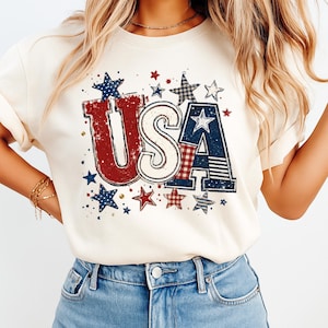 May include: A cream-colored t-shirt featuring a large graphic print of the letters USA in red, white, and blue. The design includes distressed textures, plaid and striped patterns within the letters, and various star shapes scattered around the text.