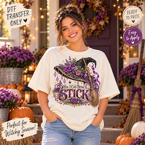 May include: A cream-colored t-shirt featuring a graphic of a black witch hat adorned with purple roses and lavender, a broomstick, and the text "Yes, I Can Drive A STICK" in purple and gold lettering with decorative flourishes.