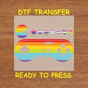 May include: A rainbow-colored video game controller with a Pac-Man design and a row of hearts in various pride flag colors. The text "DTG TRANSFER" and "READY TO PRESS" is printed on the image.