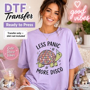 May include: A light purple t-shirt featuring a graphic of a green turtle wearing a party hat with a disco ball shell. The text reads LESS PANIC MORE DISCO, accompanied by small disco ball and musical note illustrations. This is a DTF transfer design.