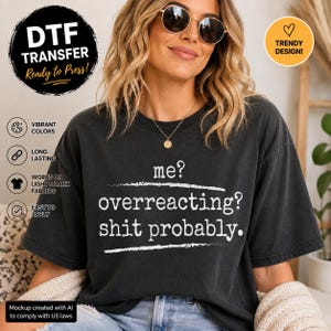 May include: A charcoal gray short-sleeve t-shirt featuring white text that reads, "me? overreacting? shit probably." The shirt has a crew neckline and a relaxed fit, with the text centered on the chest between two horizontal white lines.