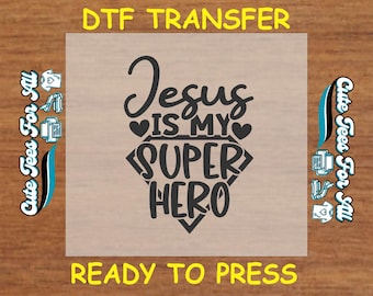 May include: A DTF transfer with the text "Jesus is my Super Hero" in black script and a diamond shape. The words "DTF TRANSFER" and "READY TO PRESS" are in yellow. The image also includes the text "Cute Tees For All".