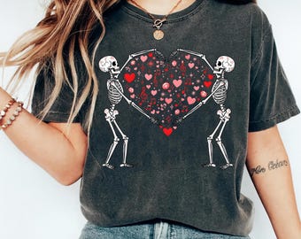 May include: Dark gray t-shirt featuring a graphic of two skeletons holding a heart filled with musical notes and hearts. The skeletons are white, and the heart design includes red and pink accents. The shirt has a vintage, worn look.